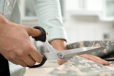 Photo of Professional tailor cutting camouflage fabric with scissors in workshop, closeup