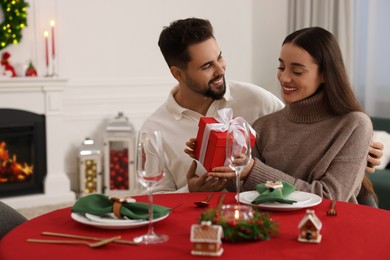 Happy young man presenting Christmas gift to his girlfriend at table indoors