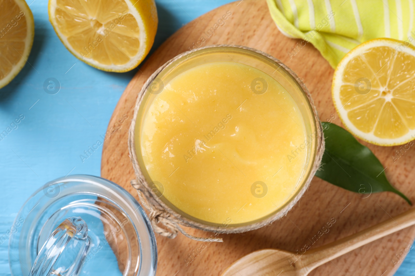 Photo of Delicious lemon curd in glass jar, fresh citrus fruits, green leaf and spoon on light blue wooden table, top view