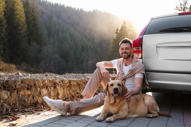 Photo of Happy man and adorable dog sitting near car in mountains. Traveling with pet