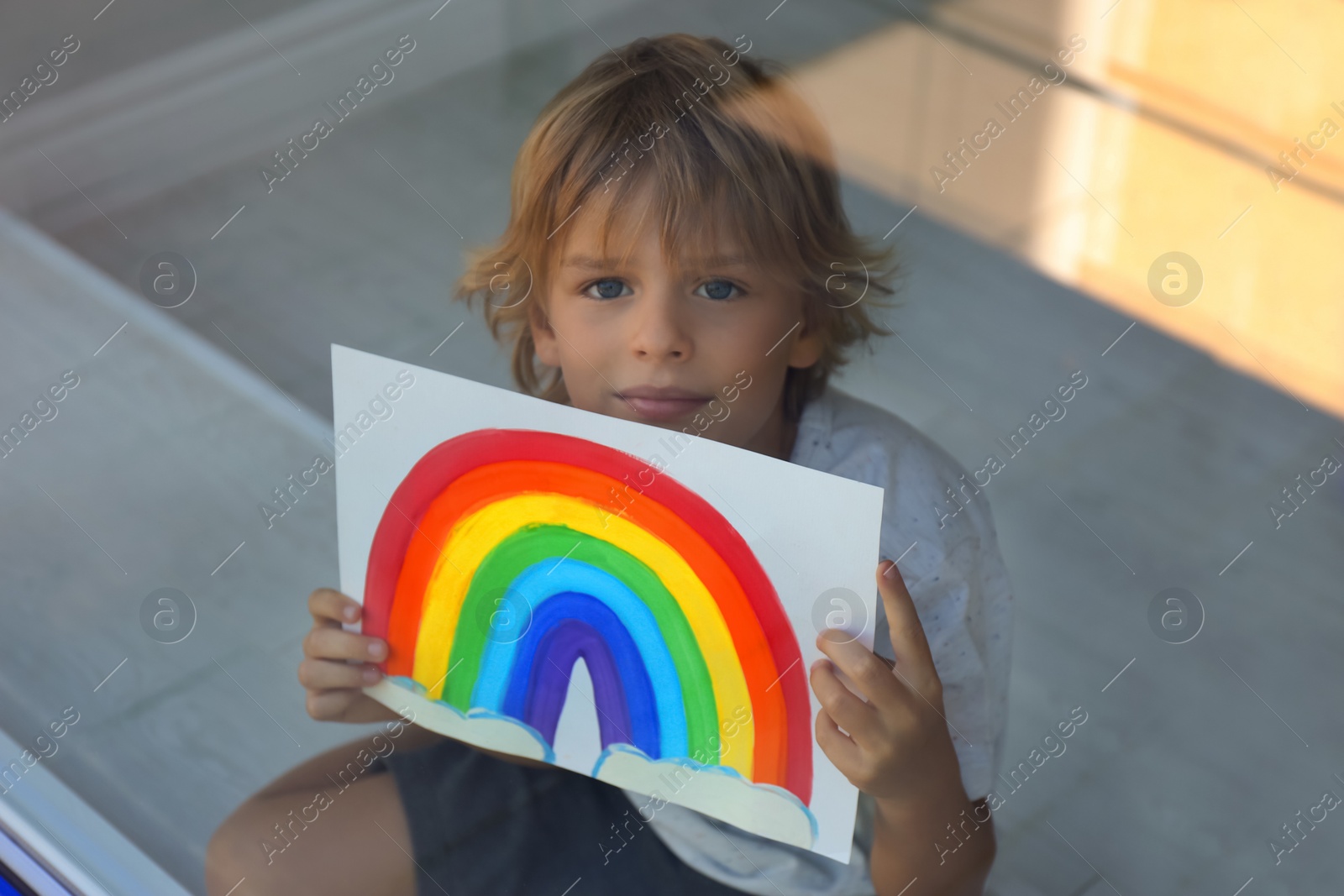 Photo of Little boy with picture of rainbow near window, view from outdoors. Stay at home concept