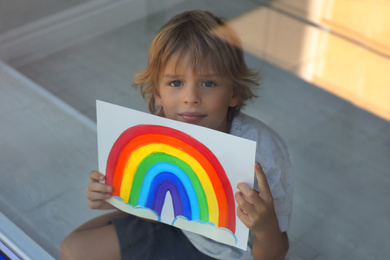 Photo of Little boy with picture of rainbow near window, view from outdoors. Stay at home concept