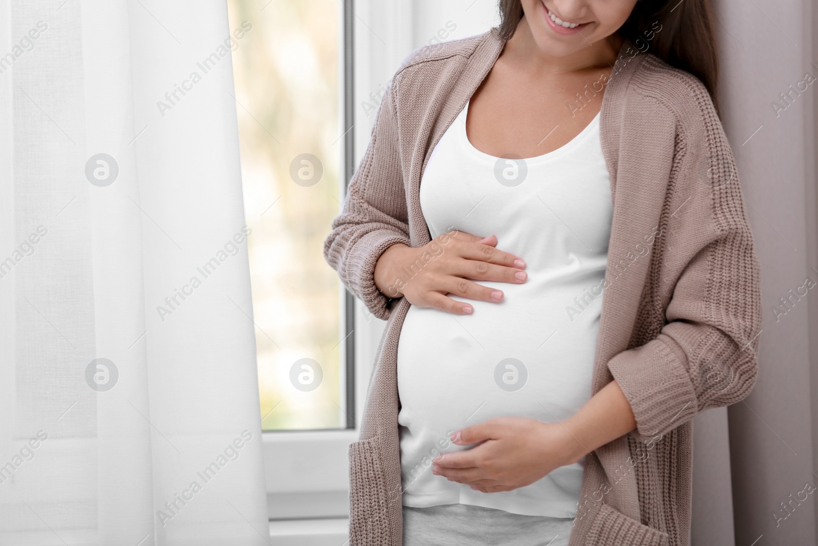 Photo of Pregnant woman standing near window at home, closeup