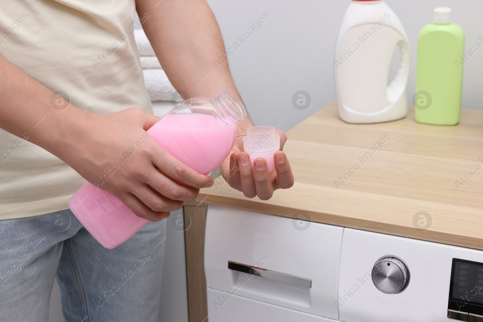 Photo of Man pouring fabric softener from bottle into cap near washing machine indoors, closeup