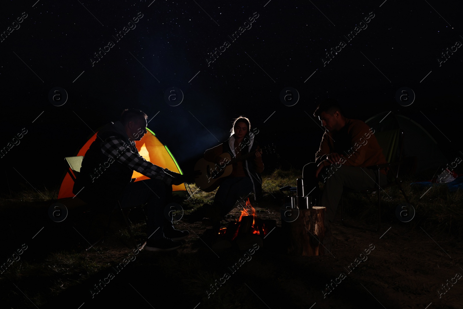 Photo of Group of friends with guitar near bonfire and camping tent outdoors at night