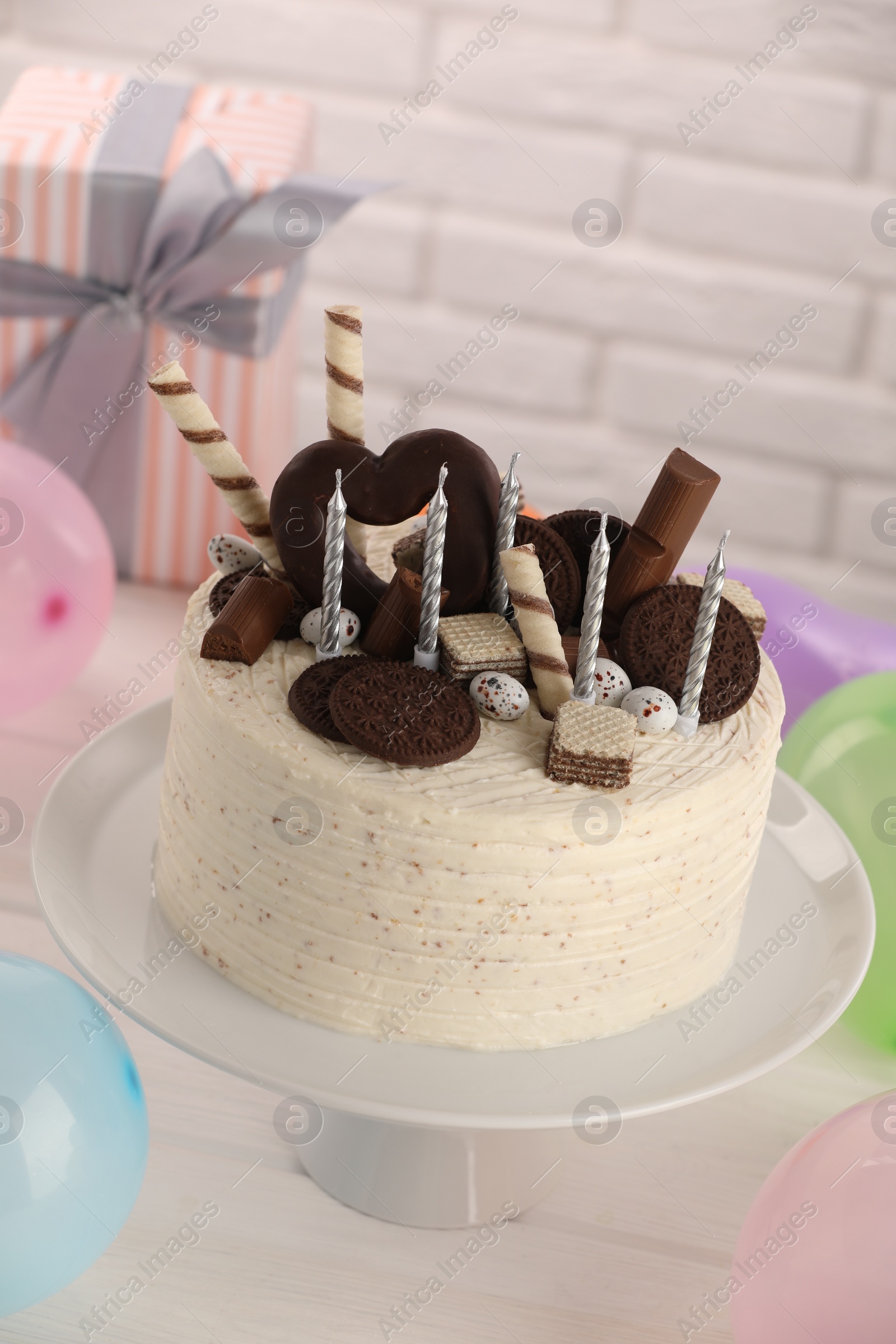 Photo of Delicious cake decorated with sweets and candles among balloons and gift box on white table, above view