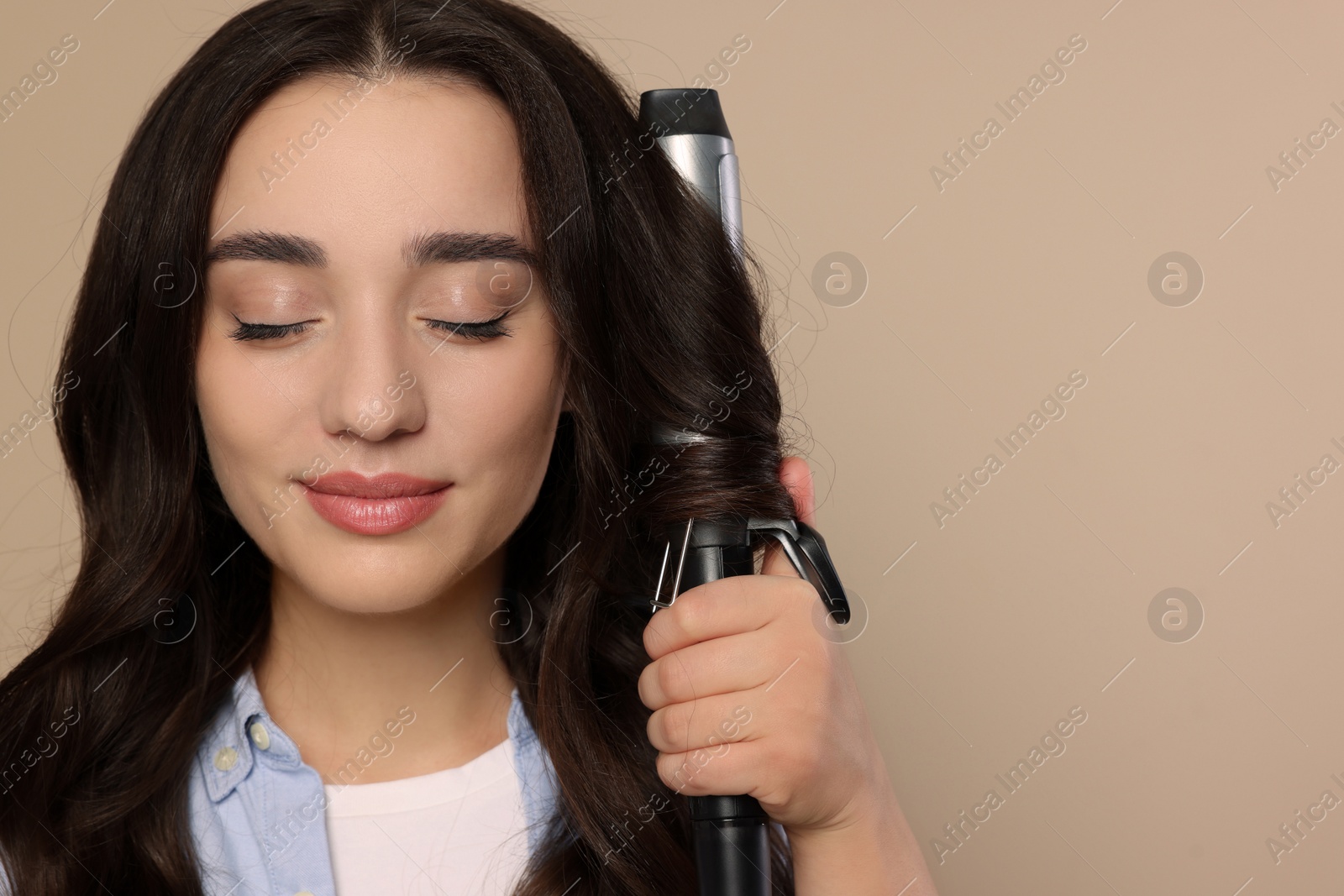 Photo of Beautiful woman using curling hair iron on beige background. Space for text
