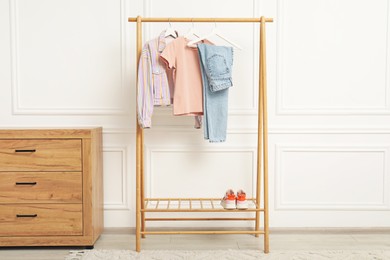 Wardrobe organization. Rack with different stylish clothes, shoes and chest of drawers near white wall indoors