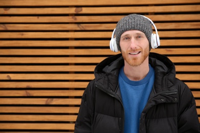 Young man listening to music with headphones against wooden wall. Space for text