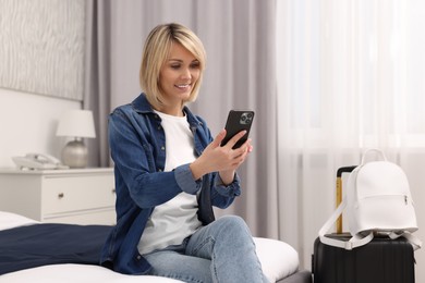 Photo of Smiling guest with smartphone relaxing on bed in stylish hotel room