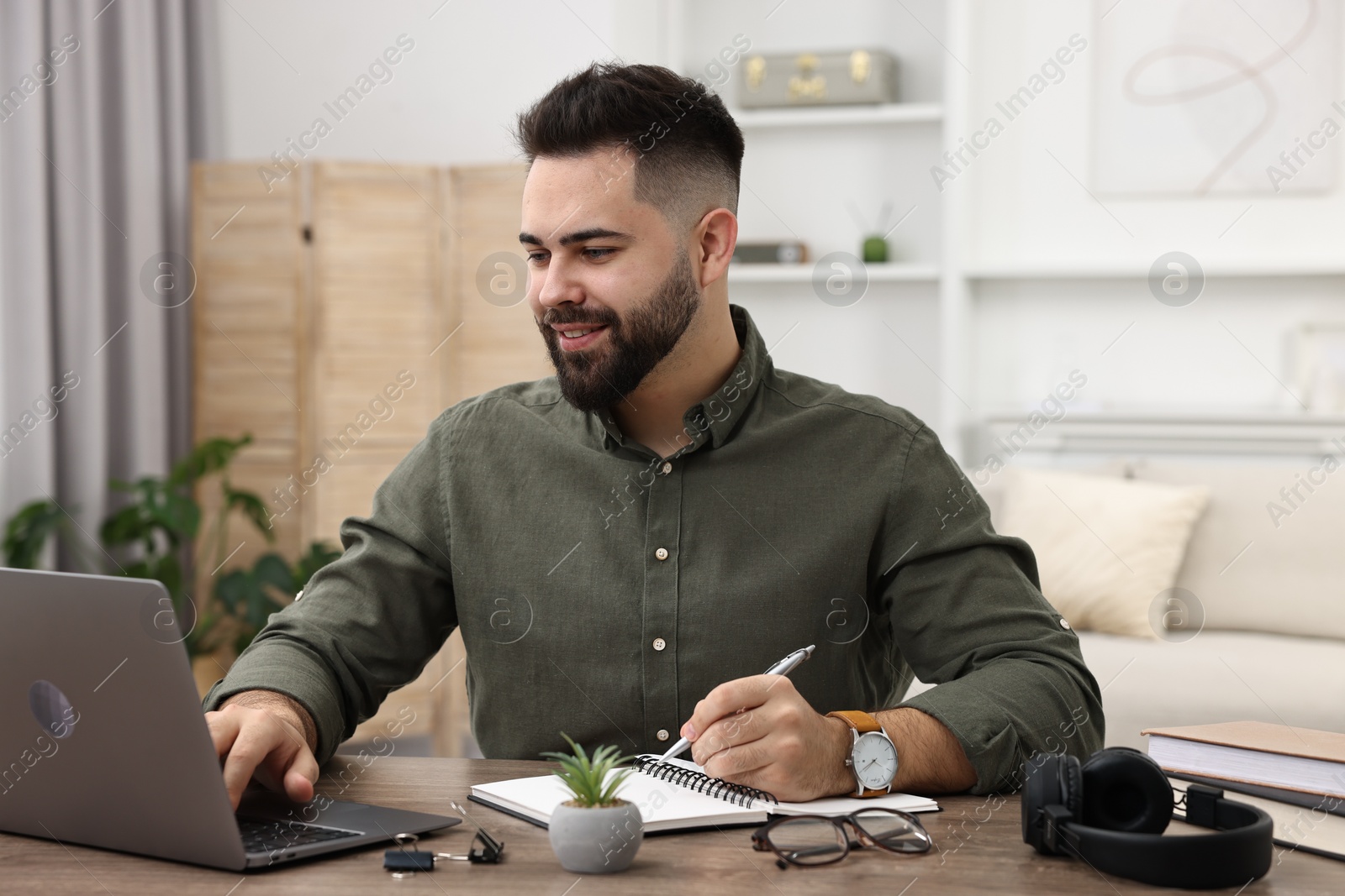 Photo of E-learning. Young man taking notes during online lesson at wooden table indoors