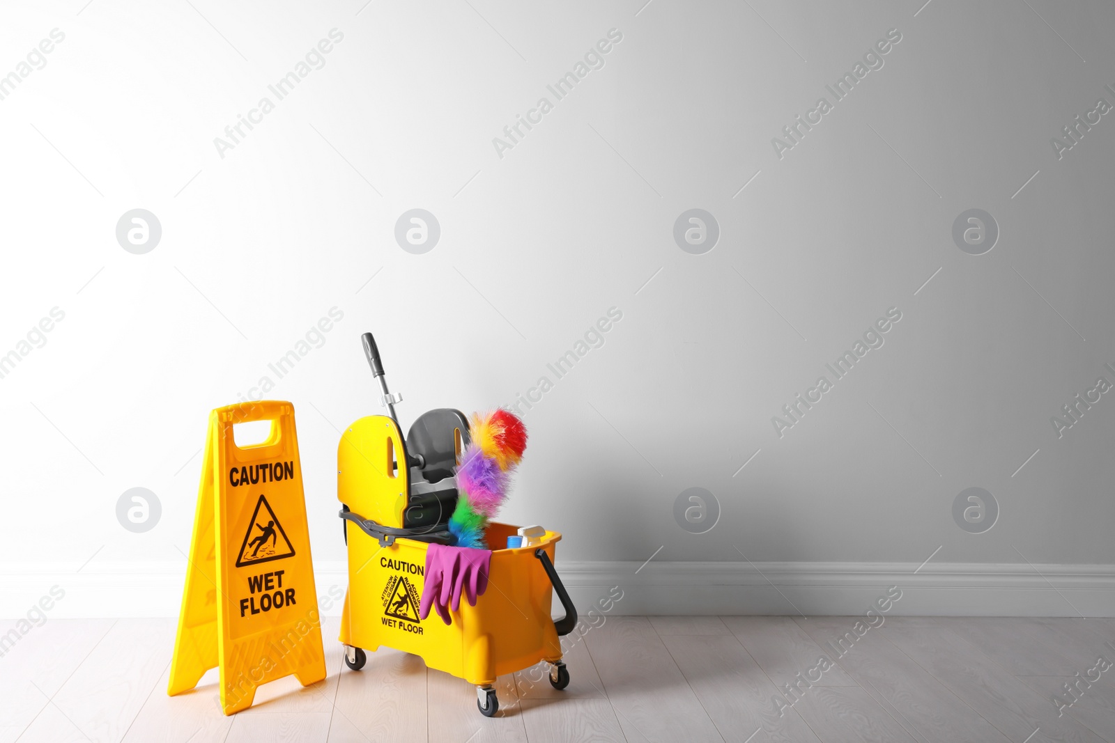 Photo of Safety sign with phrase "CAUTION WET FLOOR" and mop bucket on floor near white wall. Cleaning tools