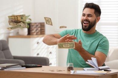 Happy young man throwing money at wooden table indoors