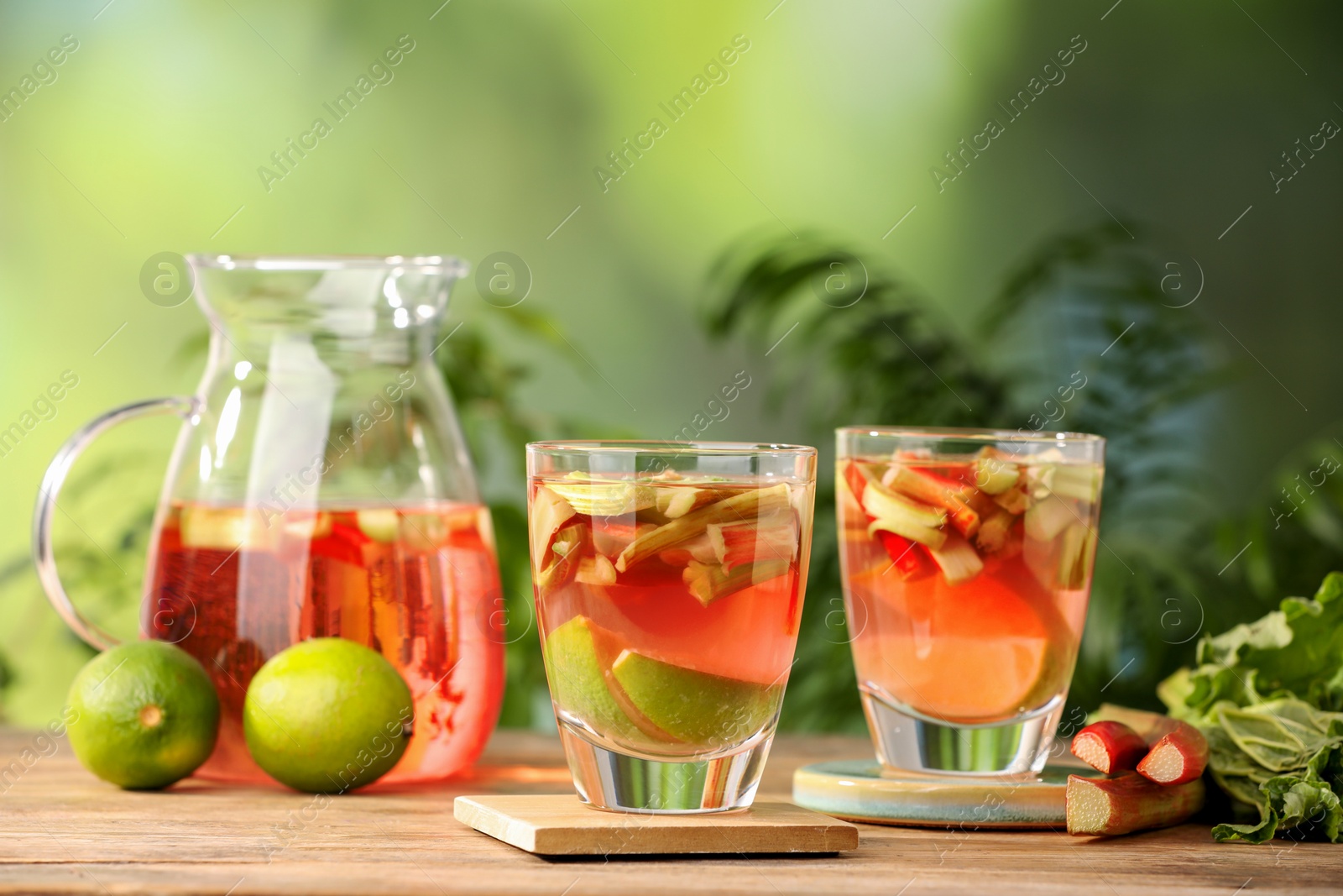 Photo of Glasses and jug of tasty rhubarb cocktail with lime fruits on wooden table outdoors