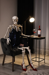 Skeleton in dress with wine sitting at table indoors