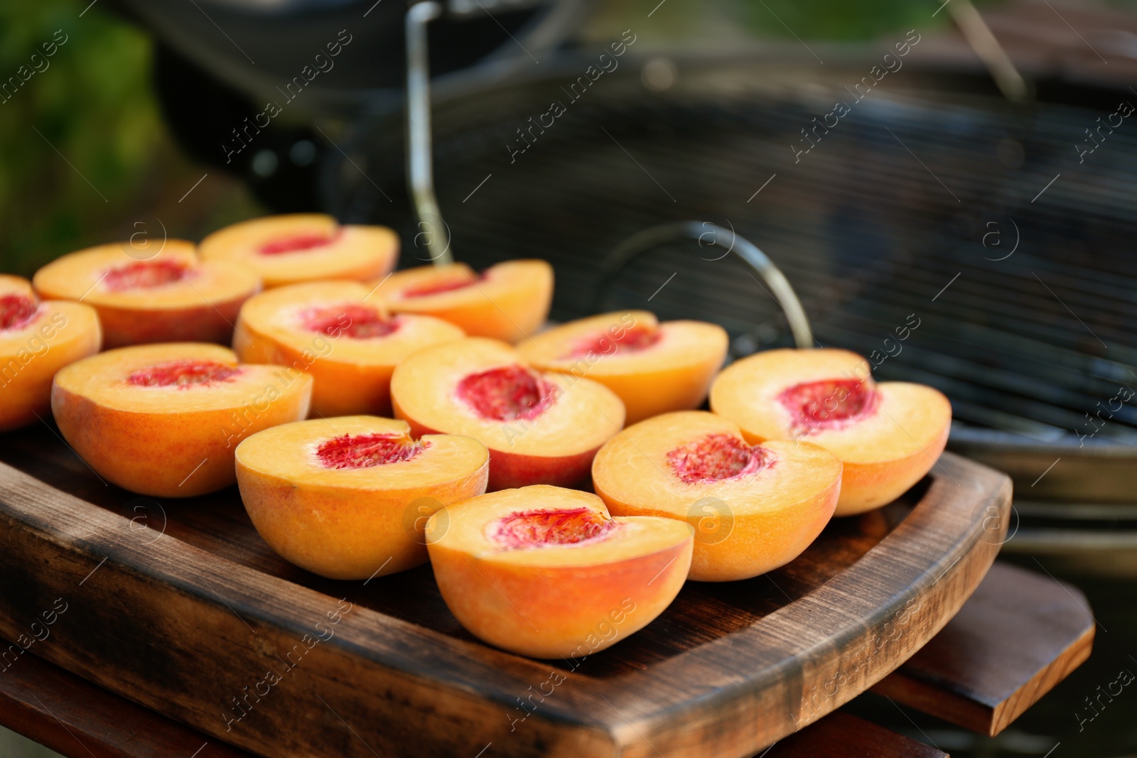 Photo of Halves of fresh peaches on wooden board near modern grill outdoors, closeup