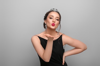 Beautiful young woman wearing luxurious tiara and blowing kiss on light grey background