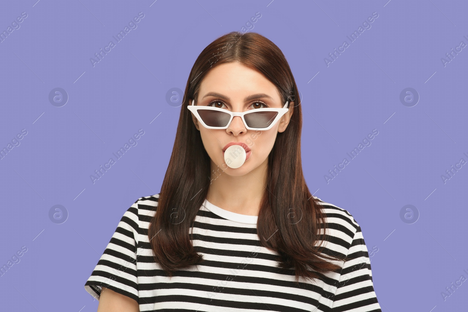 Photo of Beautiful woman in sunglasses blowing bubble gum on light purple background