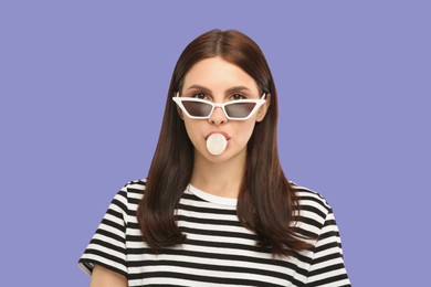 Beautiful woman in sunglasses blowing bubble gum on light purple background