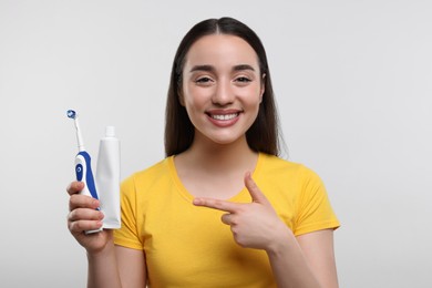 Happy young woman holding electric toothbrush and tube of toothpaste on white background