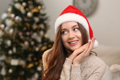 Beautiful young smiling woman in Santa hat near Christmas tree at home, space for text