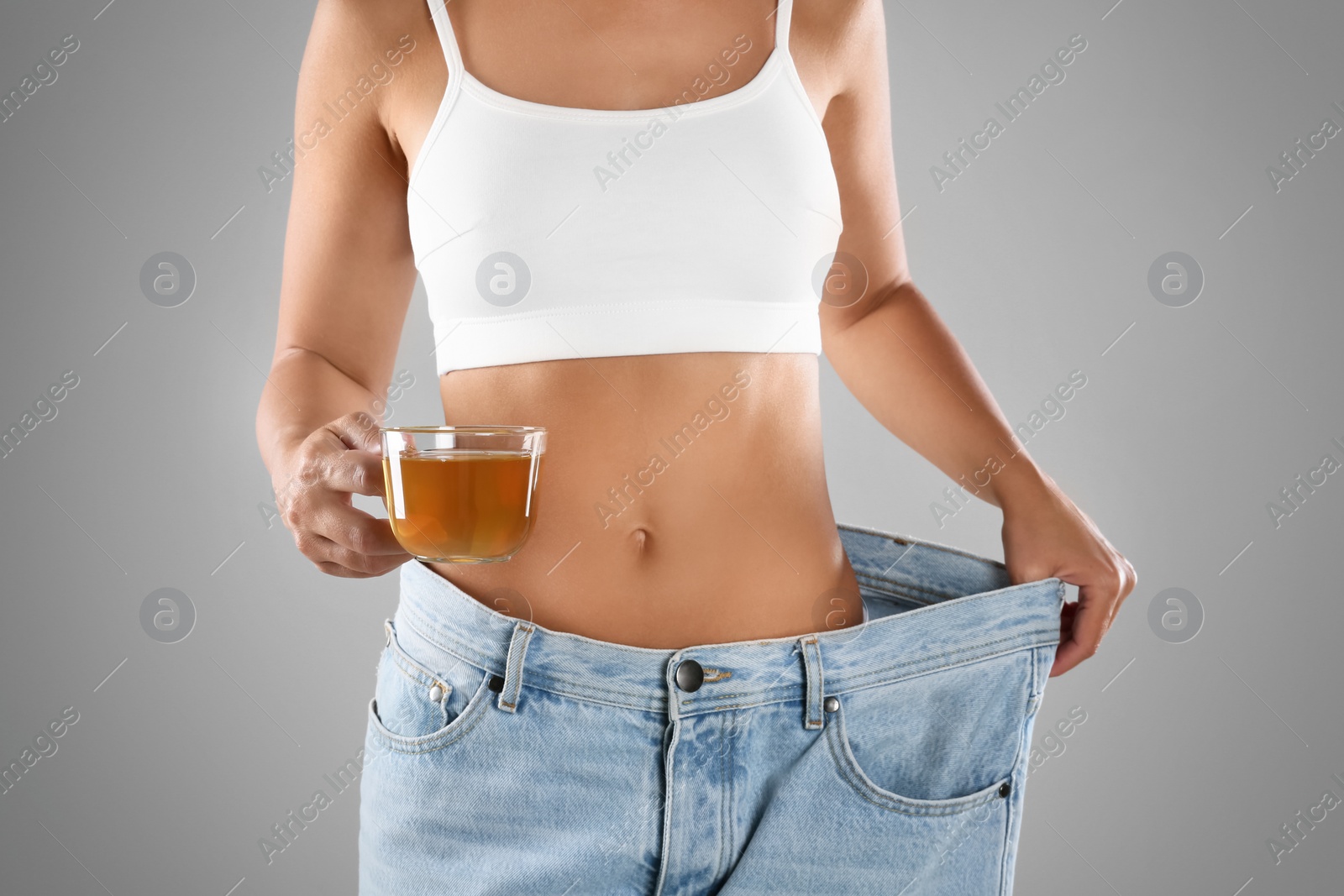 Photo of Young woman in old big jeans with cup of tea showing her diet results on grey background, closeup