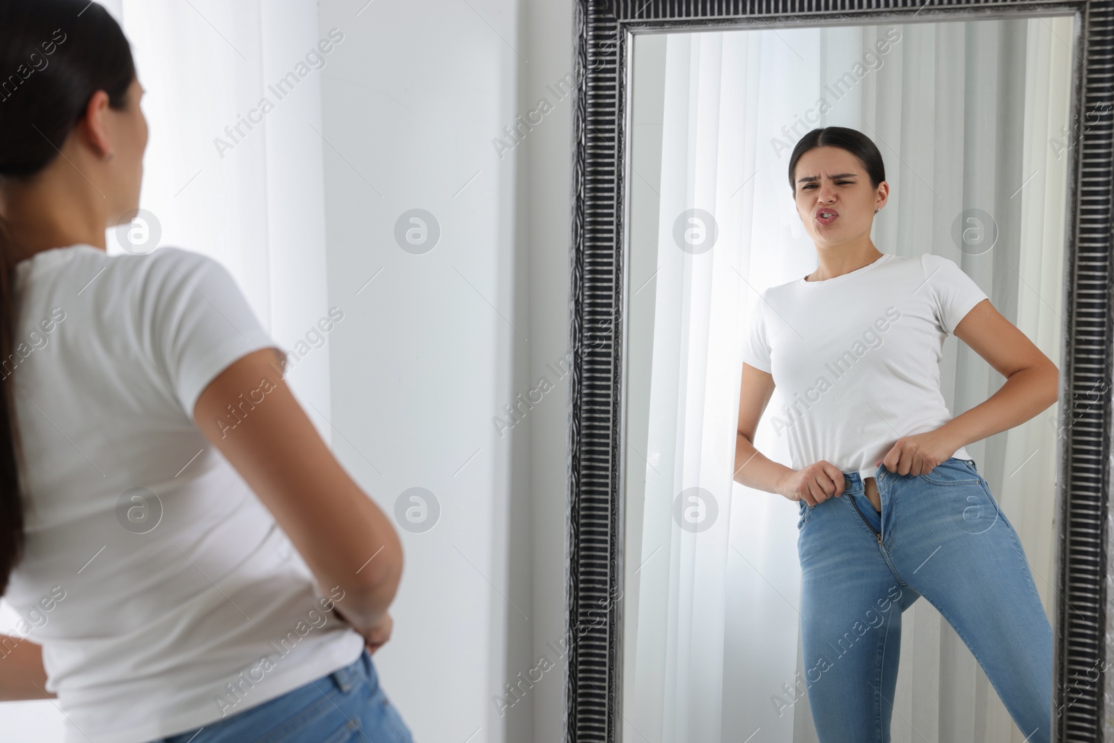 Photo of Young woman trying to put on tight jeans near mirror indoors