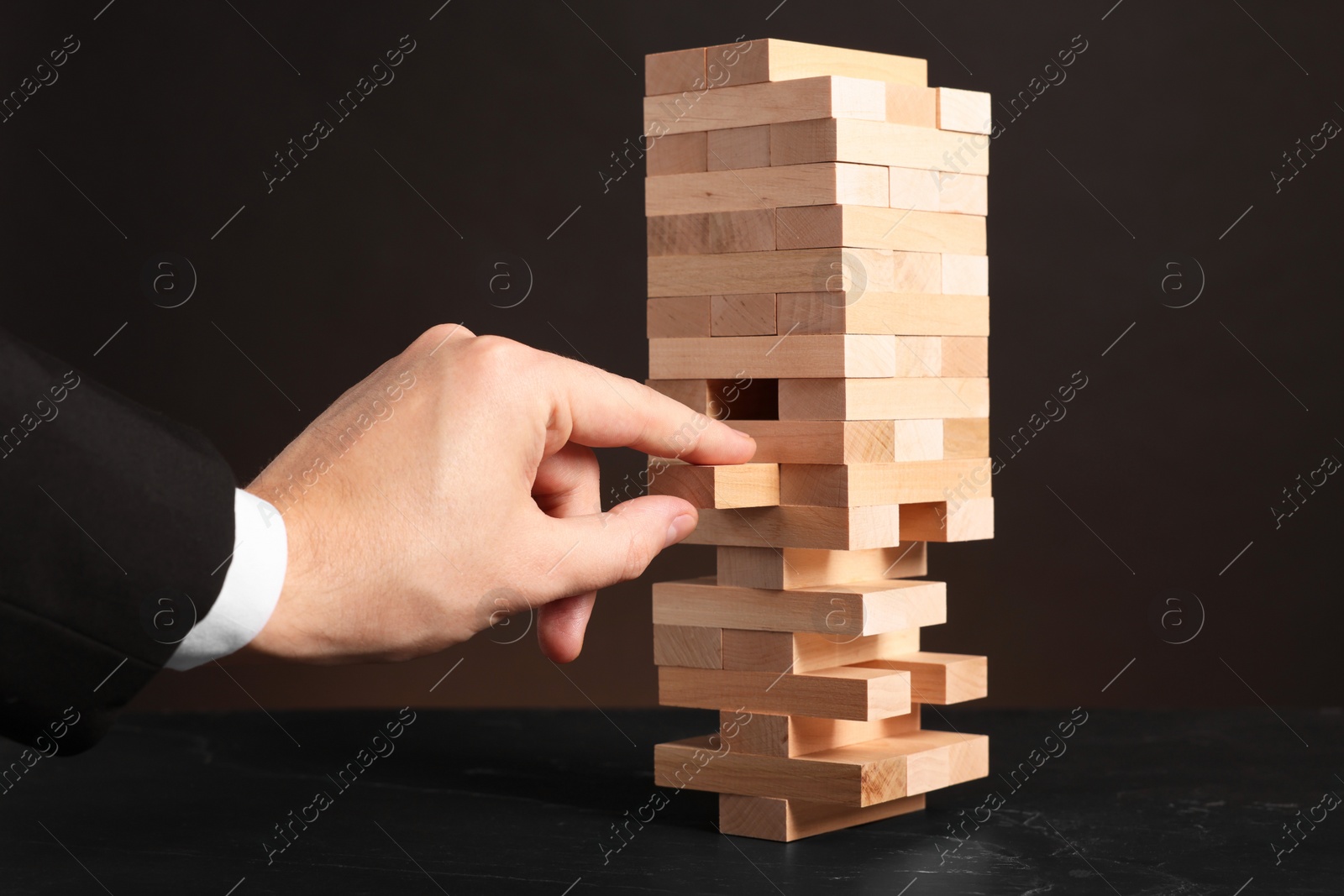 Photo of Playing Jenga. Man removing wooden block from tower at black table against dark background, closeup