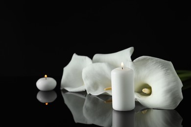 Photo of White calla lily flowers and burning candles on black mirror surface in darkness, space for text. Funeral symbols