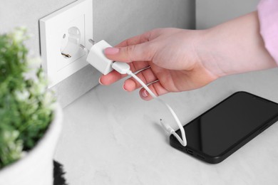 Woman plugging smartphone into power socket at white table indoors, closeup