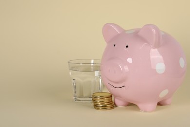 Photo of Water scarcity concept. Piggy bank, coins and glass of drink on beige background, space for text