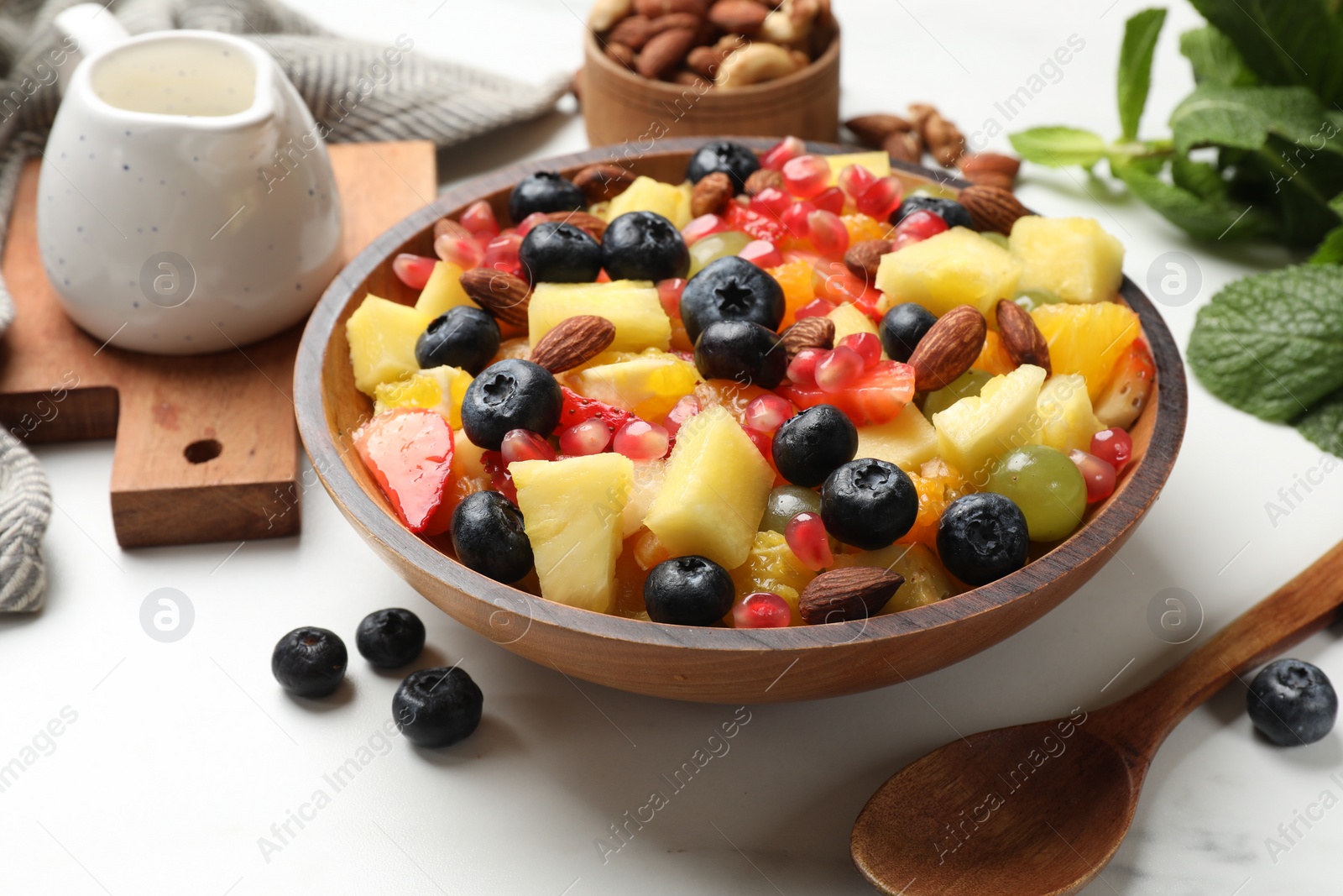 Photo of Delicious fruit salad in bowl, berries, fresh mint and nuts on white marble table