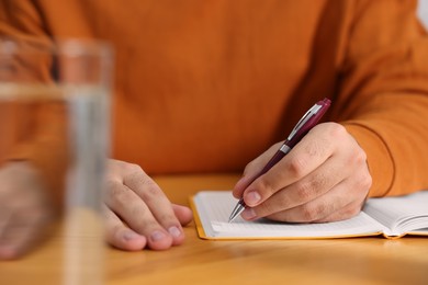 Photo of Young man writing in notebook at wooden table, closeup