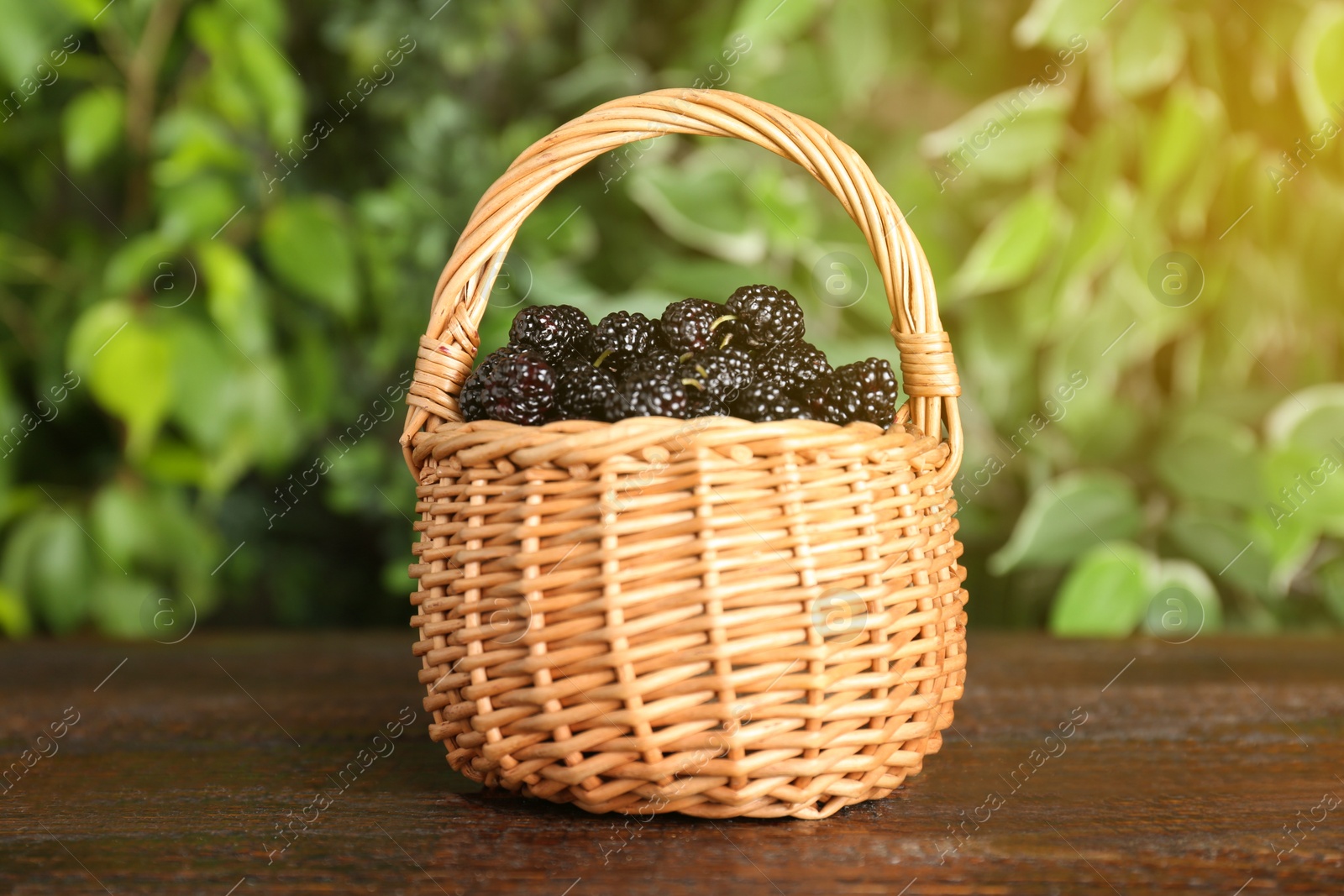 Photo of Ripe black mulberries in wicker basket on wooden table against blurred background