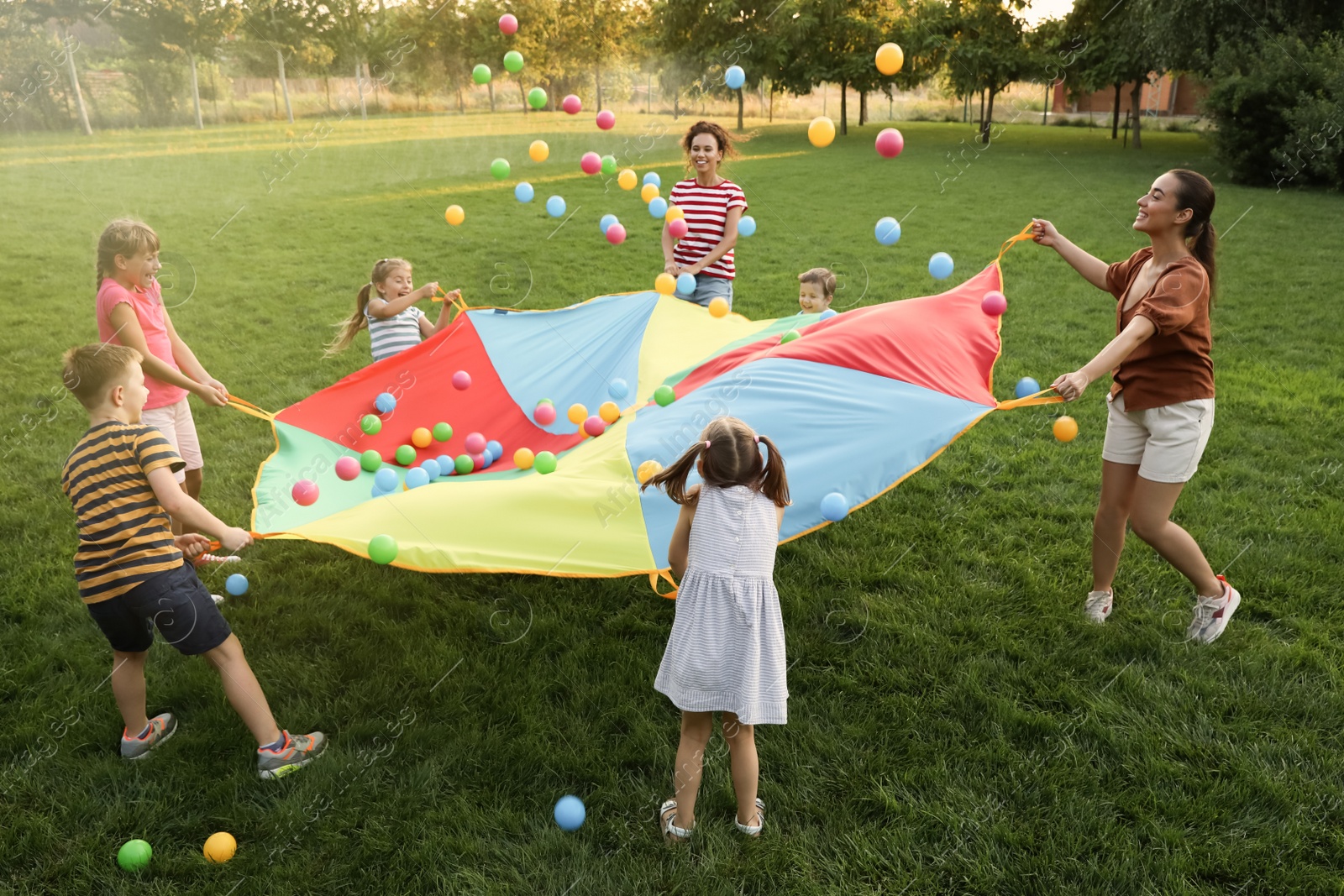 Photo of Group of children and teachers playing with rainbow playground parachute on green grass. Summer camp activity