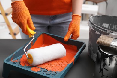 Woman taking orange paint with roller from tray at grey table indoors, closeup