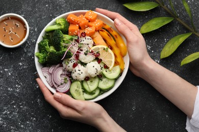 Photo of Woman holding bowl with many different vegetables and rice at grey textured table, top view. Vegan diet
