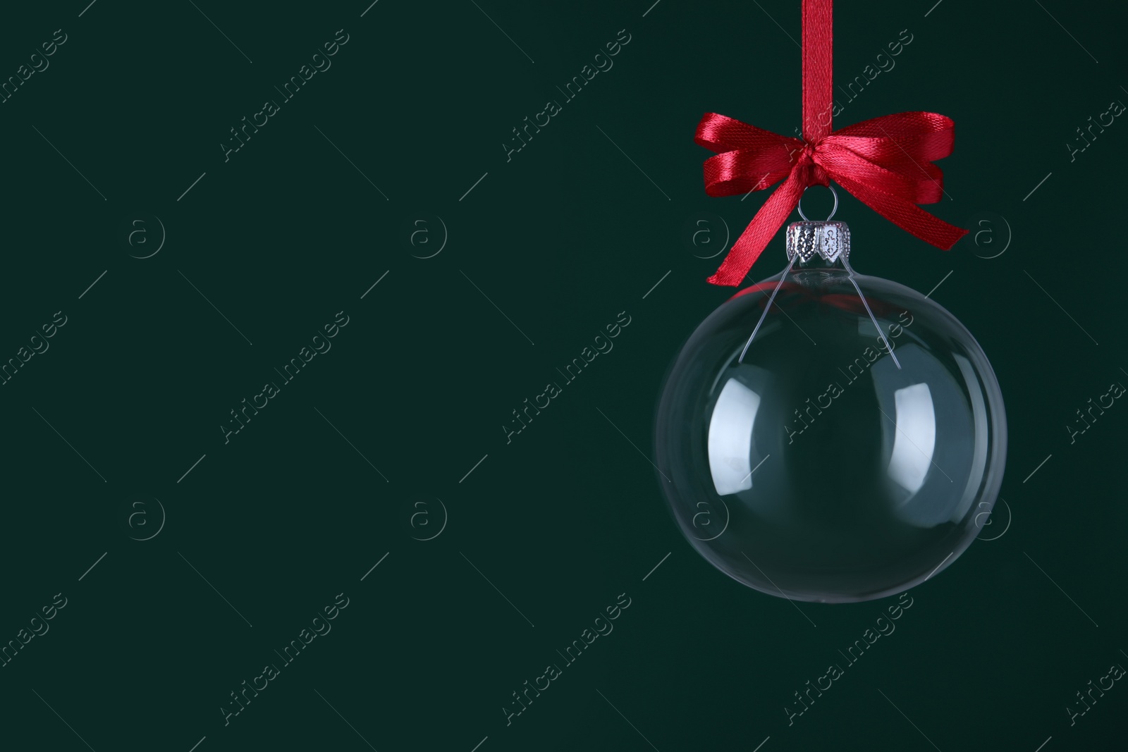 Photo of Transparent glass Christmas ball with red ribbon and bow against dark green background. Space for text