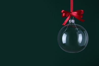 Photo of Transparent glass Christmas ball with red ribbon and bow against dark green background. Space for text