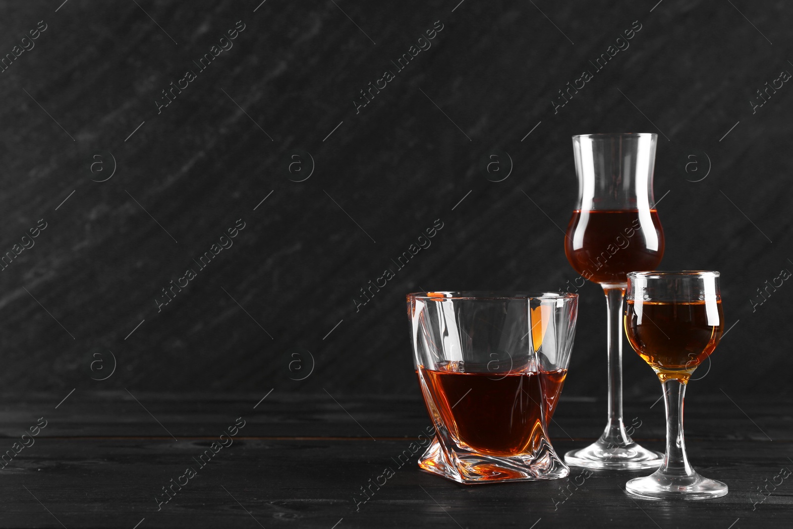 Photo of Different delicious liqueurs in glasses on black wooden table, space for text