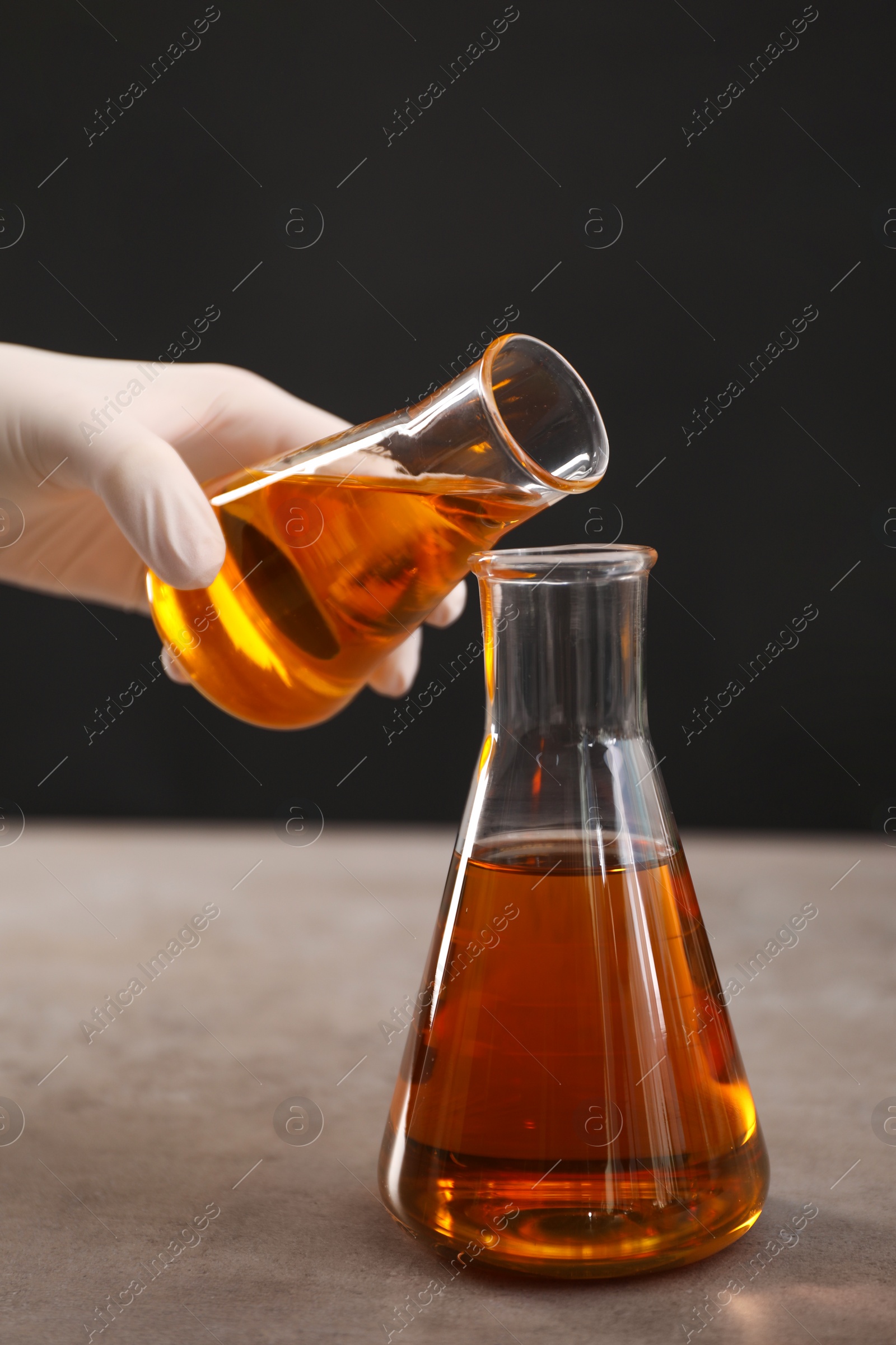 Photo of Woman pouring yellow crude oil into flask at grey table against dark background, closeup