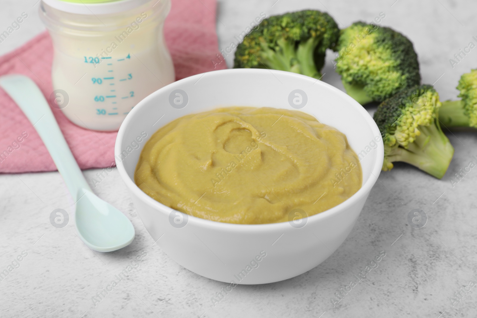 Photo of Bowl with healthy baby food, broccoli, spoon and bottle of milk on white table, closeup