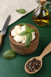 Tasty mozzarella balls and basil leaves in bowl on green wooden table