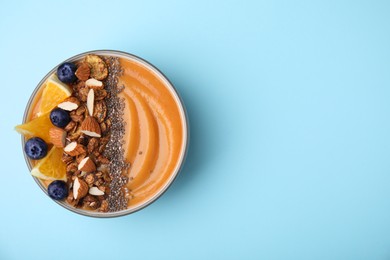 Photo of Bowl of delicious fruit smoothie with fresh orange slices, blueberries and granola on light blue background, top view. Space for text