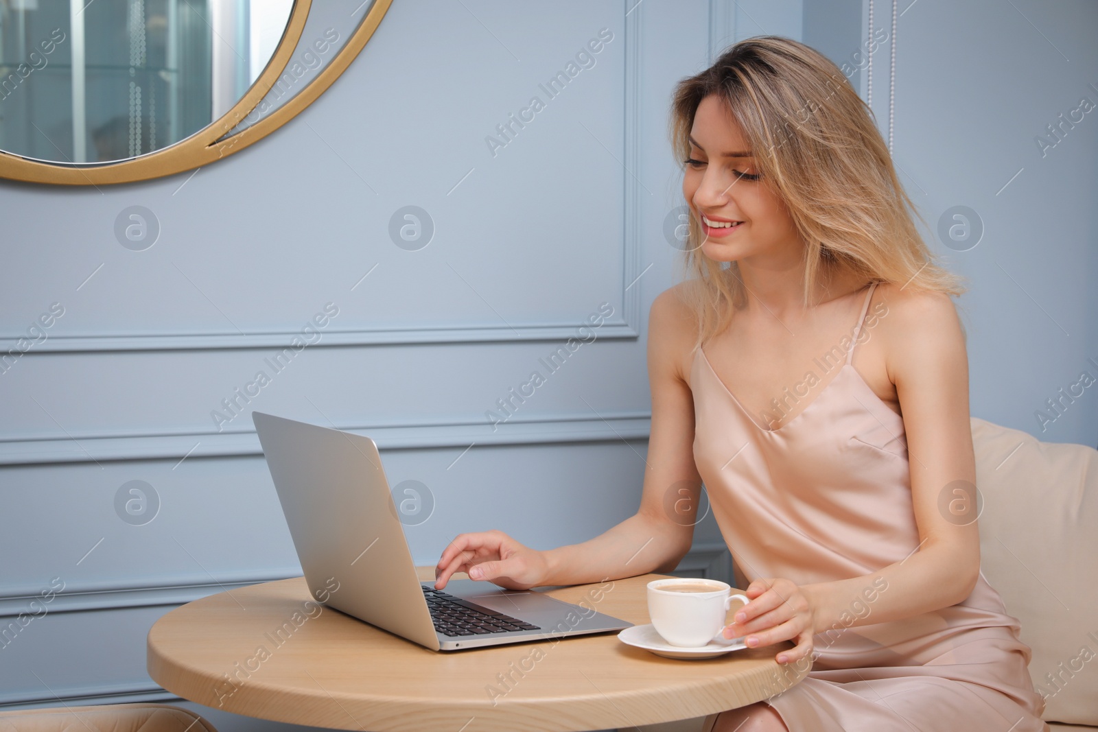 Photo of Happy young woman with cup of coffee and laptop at table indoors in morning