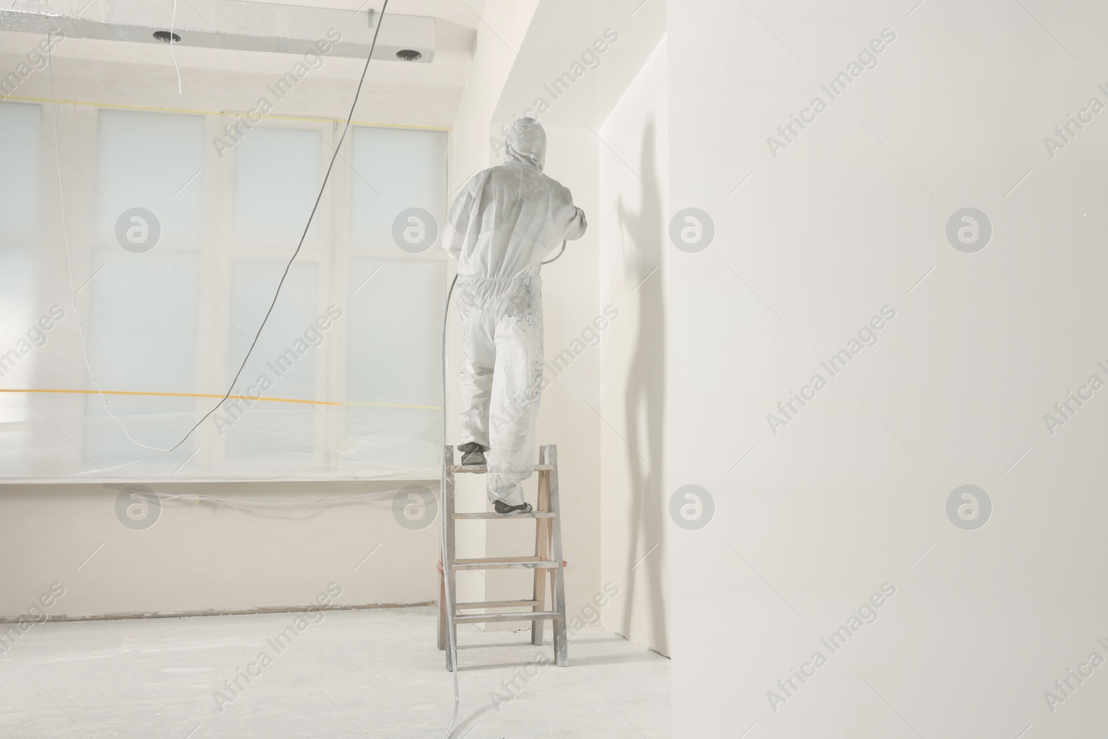 Photo of Decorator in protective overalls painting wall with spray gun indoors