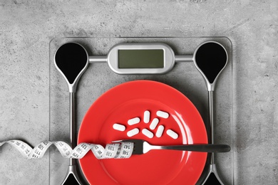 Scales with plate of weight loss pills, fork and measuring tape on gray background, top view