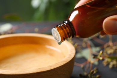 Photo of Woman dripping eucalyptus essential oil from bottle into bowl on blurred background, closeup