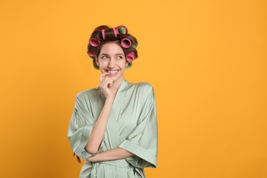 Photo of Happy young woman in silk bathrobe with hair curlers on yellow background. Space for text