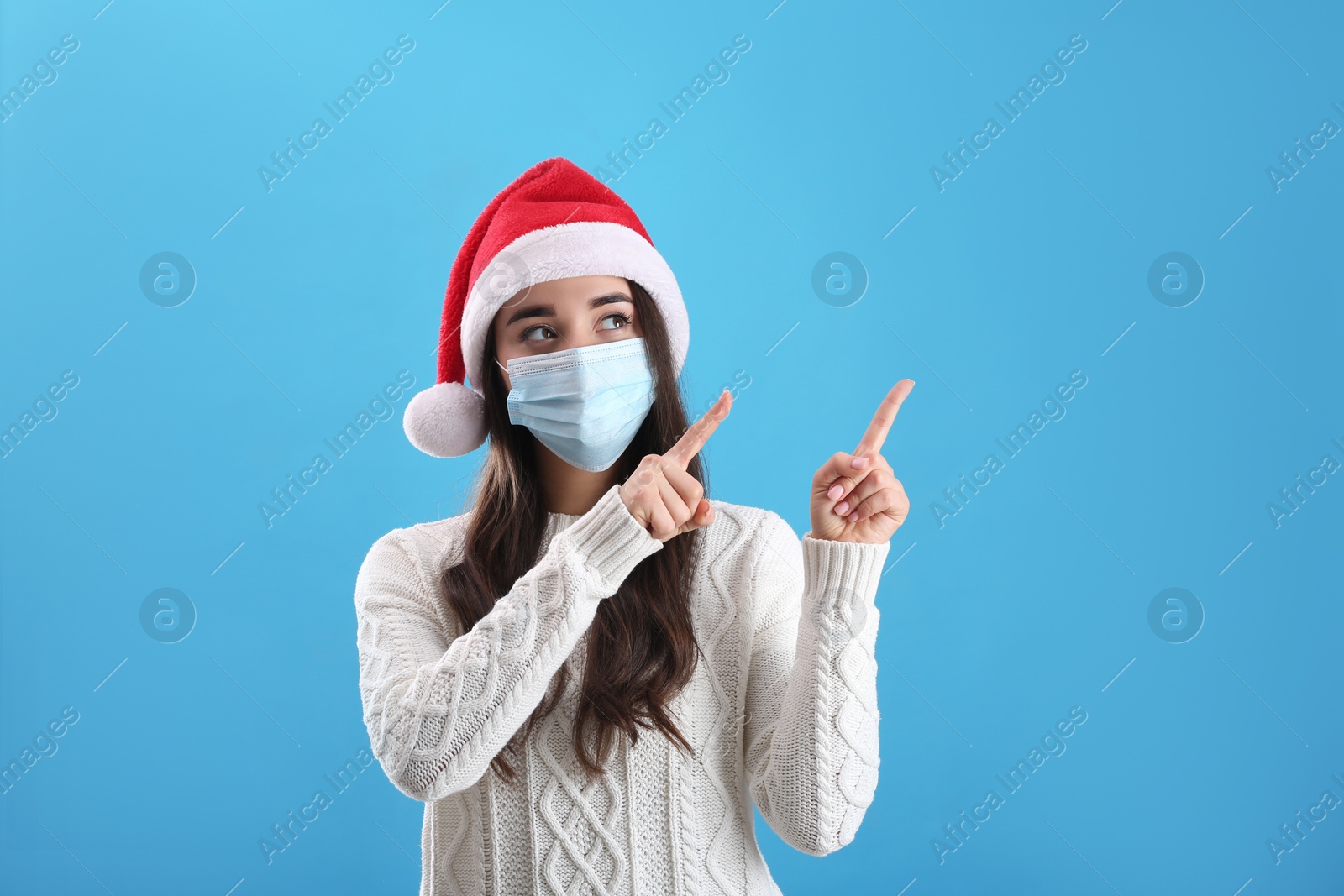 Photo of Beautiful woman wearing Santa Claus hat and medical mask on light blue background